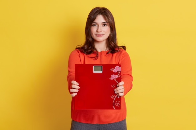 Dark haired female wearing orange casual jumper showing floor scales, isolated over yellow wall.