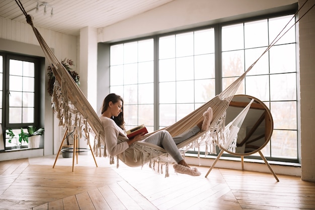Темноволосая девушка в брюках, свитере и теплых тапочках читает книгу, лежа в гамаке в уютной комнате с деревянным полом, панорамными окнами и круглым зеркалом на полу. .