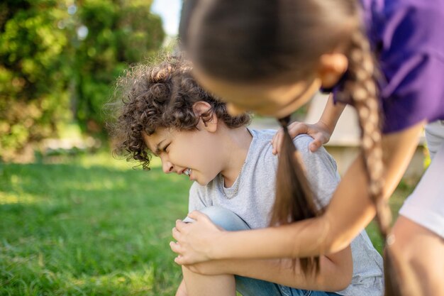 転倒後の膝の痛みに苦しんでいる白人の少年に寄りかかっている黒髪の少女