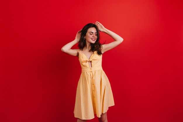 Dark-haired good-humored girl relaxed posing on red wall. Woman in bright summer outfit smiling.