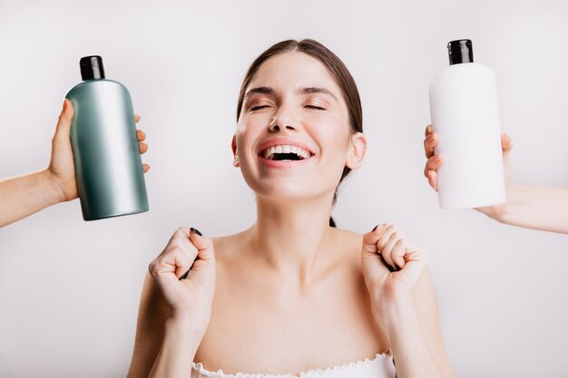 Dark-haired lady happily smiles, posing on isolated wall with bottles of shampoos.