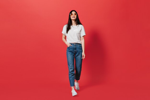 Dark-haired lady in orange glasses, dressed in jeans and top moves on red wall