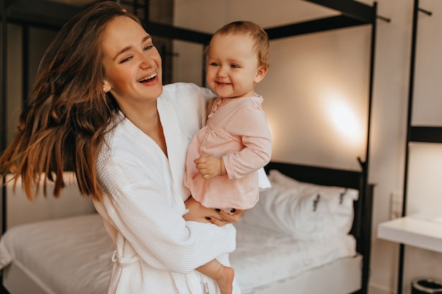 Dark-haired lady in white bathrobe and her little daughter laugh sincerely while playing in bright bedroom.