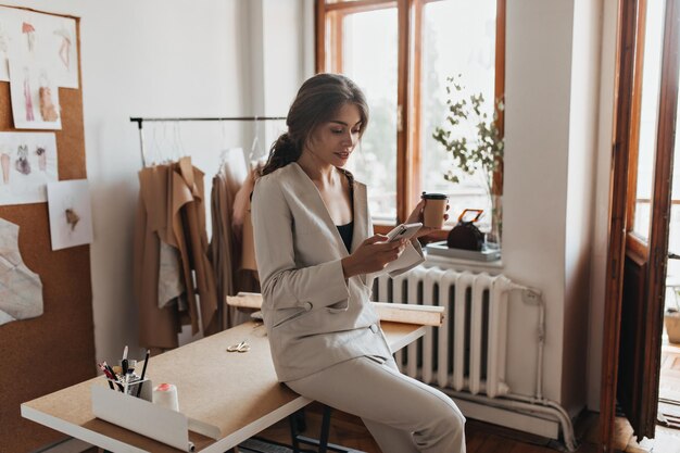 Dark haired lady in white suit holding coffee and phone Charming woman in linen pants and beige blouse chatting in cellphone and drinking tea