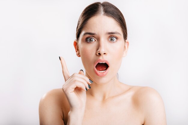 Dark-haired lady with green eyes posing on white wall. Young woman without makeup remembered shocking news.