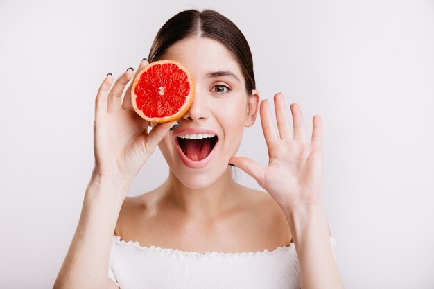 Dark-haired lady with healthy skin with smile, covering her eyes with red grapefruit.