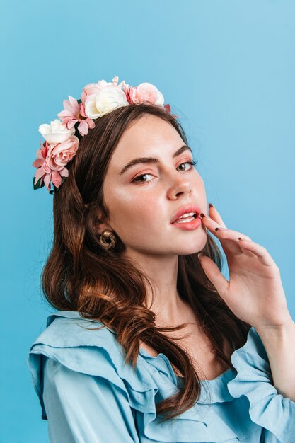 Dark-haired lady with plump lips . Green-eyed girl with long eyelashes and in crown of flowers posing on blue wall.