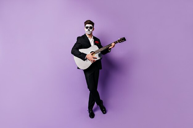 Dark-haired man in elegant suit and painted skull-shaped face plays guitar, looking into camera with deadpan look.