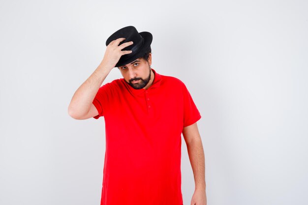Dark-haired man in red t-shirt adjusting hat , front view.