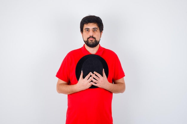 Dark-haired man in red t-shirt holding hat on chest and looking calm , front view.