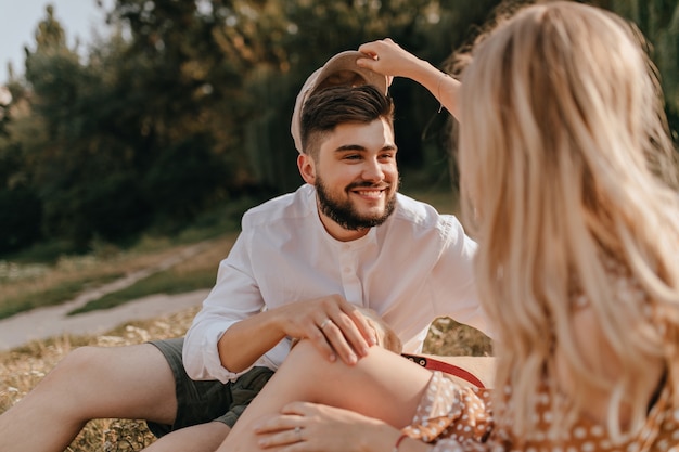 笑顔の黒髪の男は彼の帽子をかぶっている彼のガールフレンドを見ます。植物園で休んでいるカップル。