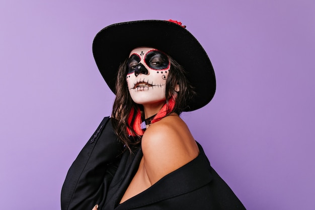 Dark-haired passionate lady with Halloween face art, holding black hat.