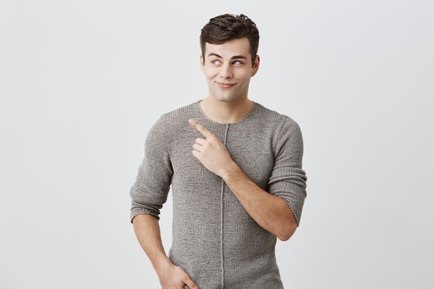 Dark-haired stylish guy in sweater looking with his blue eyes aside pointing with index finger at copy space advertising something. Man posing against wall with copy space for text or promotion