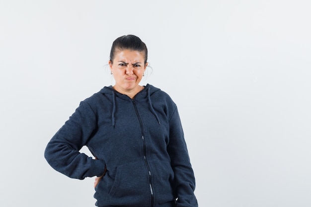 Dark-haired woman holding hand on her waist while souring face in jacket and looking displeased. front view.