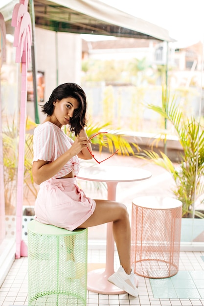 Dark-haired woman holding sunglasses in outdoor cafe. Playful young woman posing in summer morning.