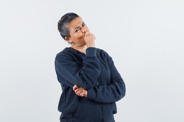 Dark-haired woman in jacket holding hand on mouth and looking pensive 