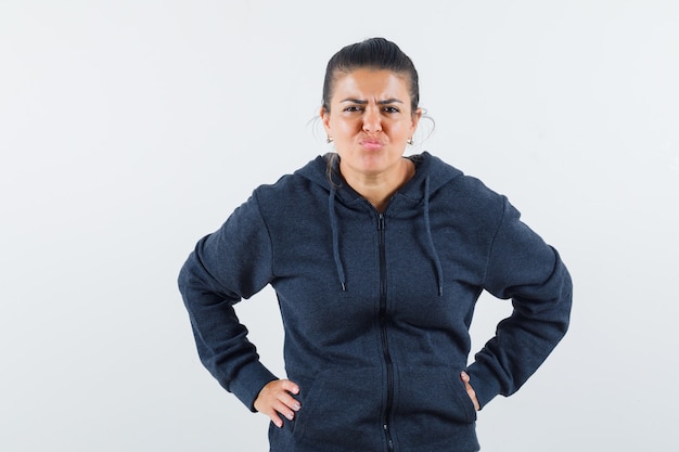 Dark-haired woman in jacket holding hands on waist and looking dissatisfied 