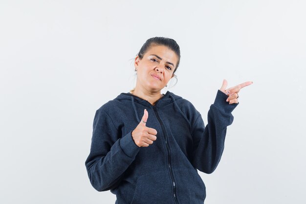 Dark-haired woman pointing aside while showing thumb up in jacket front view.