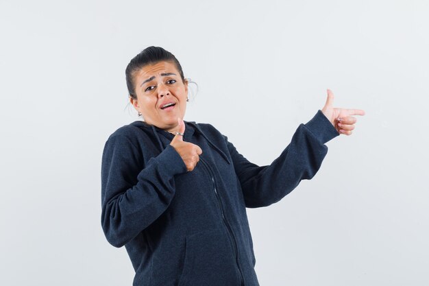 Dark-haired woman pointing aside while showing thumb up in jacket and looking suggestive 