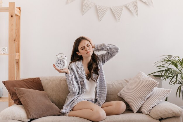 Dark-haired woman posing with alarm clock sitting on couch