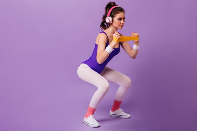Dark-haired woman in purple tracksuit and white sneakers squatting with band for sports