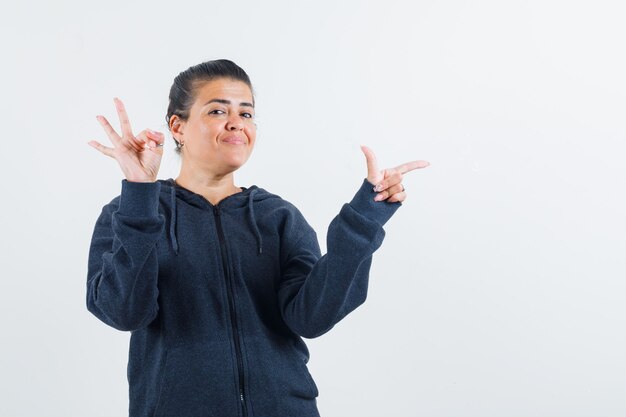 Dark-haired woman showing ok gesture while pointing aside in jacket and looking glad 