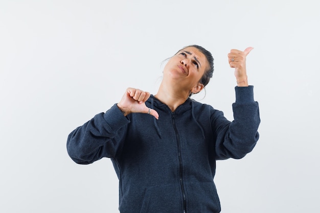 Dark-haired woman showing thumb up and down in jacket and looking perplexed 