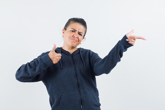 Dark-haired woman showing thumb up while pointing aside in jacket and looking anxious 