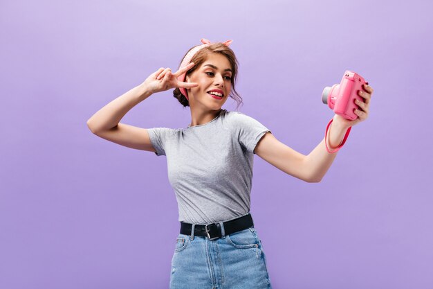 Dark-haired woman shows peace sign and holds camera. Cool cute girl in grey t-shirt and denim skirt with black belt posing.