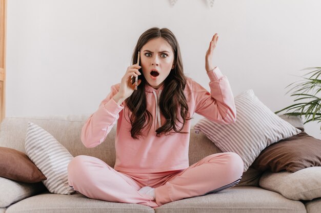 Dark-haired woman sitting on light couch surrounded by soft pillows leads unpleasant conversation on phone and waves her hands with displeasure