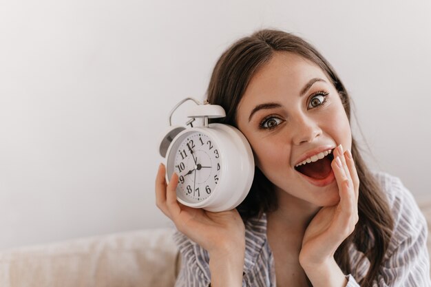 Dark-haired woman stares in surprise at front with her mouth open and keeps alarm clock