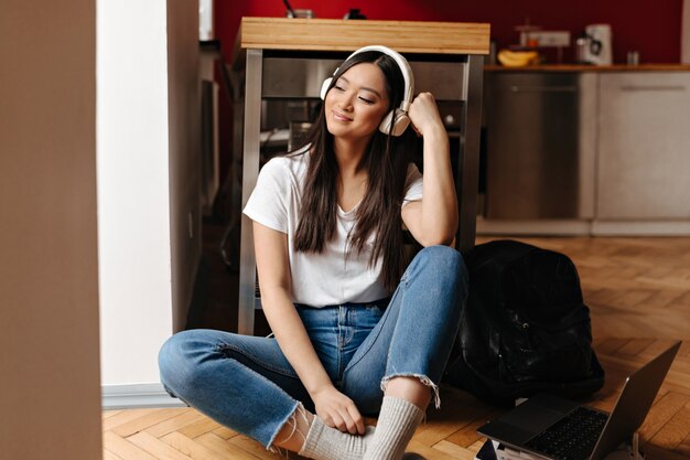 Dark-haired woman in white top and denim pants enjoys music in headphones