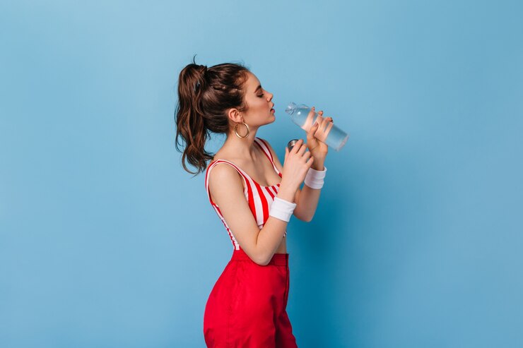 Free Photo dark-haired woman with massive earrings in red top and trousers drinking water on blue wall