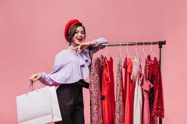 Dark-haired woman with red lipstick smiles, leans on stand with clothes and holds package on pink background.