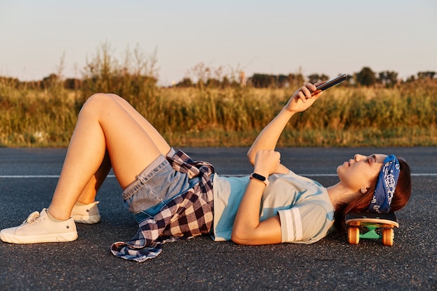 Free photo dark haired young beautiful lady lay down on the asphalt road outdoor with hand on skateboard, using a mobile phone to take selfie or cheking social networks.