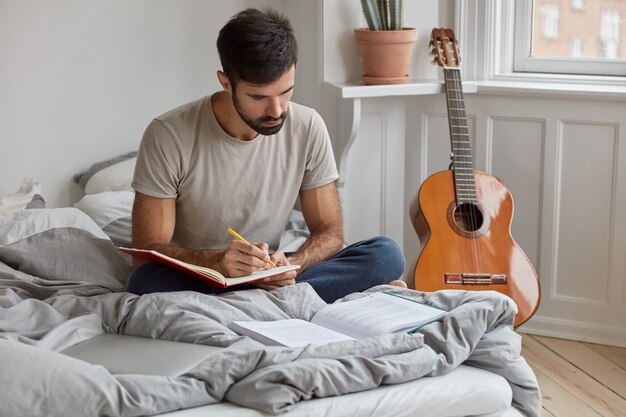 Dark haired young man in t shirt, writes information from book into notebook