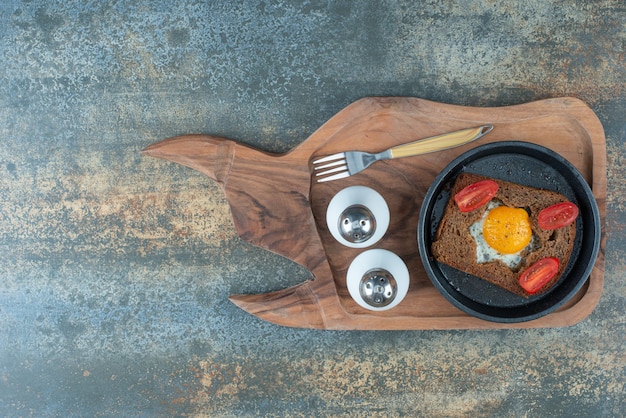 A dark pan with fried egg and slices of brown bread on wooden board