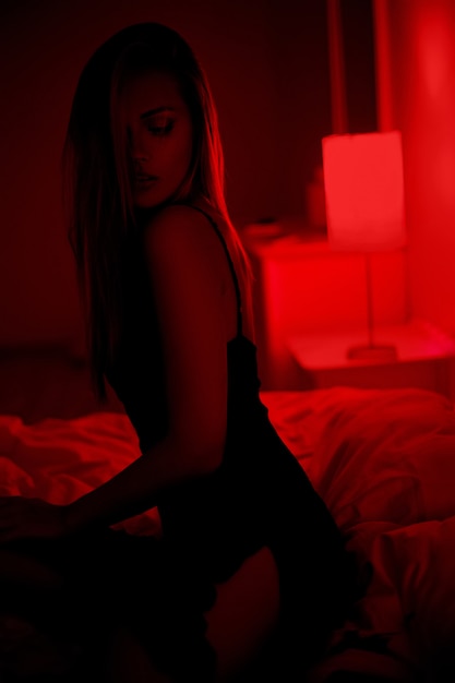 Dark photo of sexy beautiful brunette woman sitting on bed in black dress posing in red interior