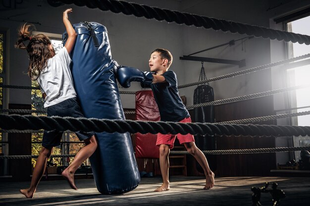 Dark photo shoot of kids training with big punching bag at boxing studio.