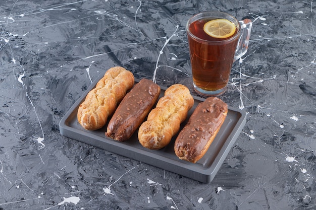 Dark plate of chocolate eclairs and glass of tea with lemon on marble surface.