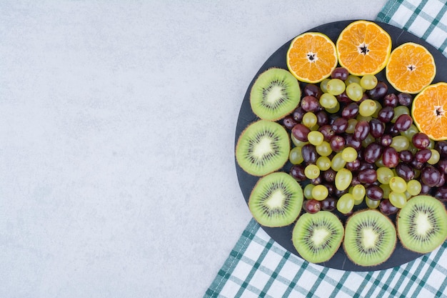 A dark plate full of grapes, kiwi and orange on tablecloth. high quality photo