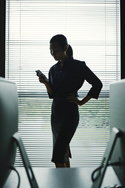 Темный силуэт женщины с смартфон стоял в офисе у окна с жалюзи