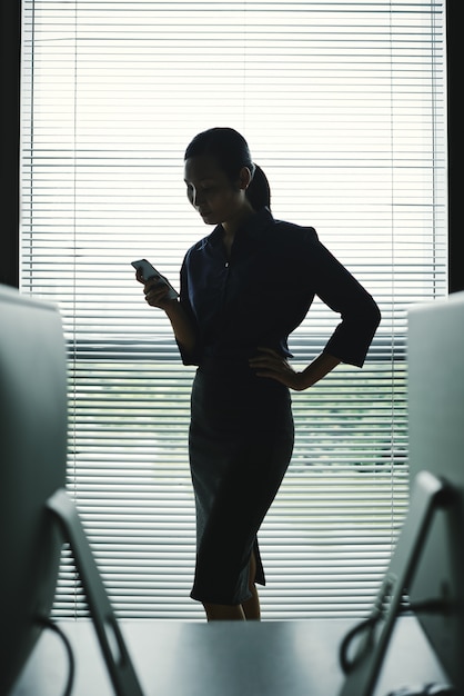 ブラインドと窓のオフィスでスマートフォンの立っている女性の暗いシルエット