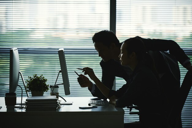 Dark silhouettes of colleagues pointing at computer screen in office against window