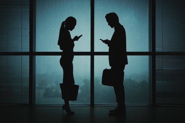 Free photo dark silhouettes of man and woman in business attire standing by window at night with smartphones