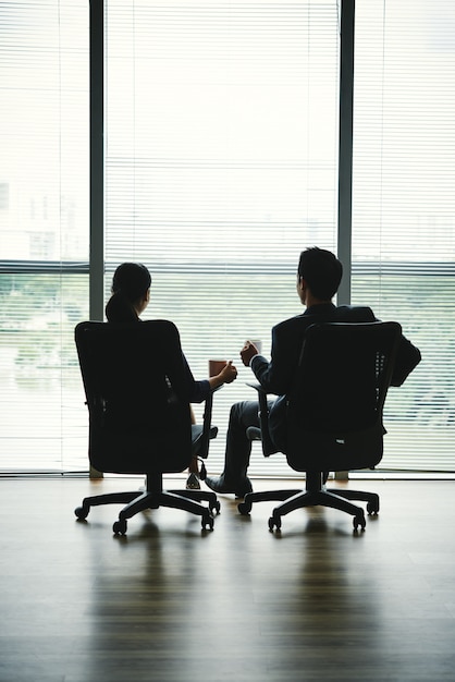 Dark silhouettes of man and woman sitting with mugs in office chairs in front of window