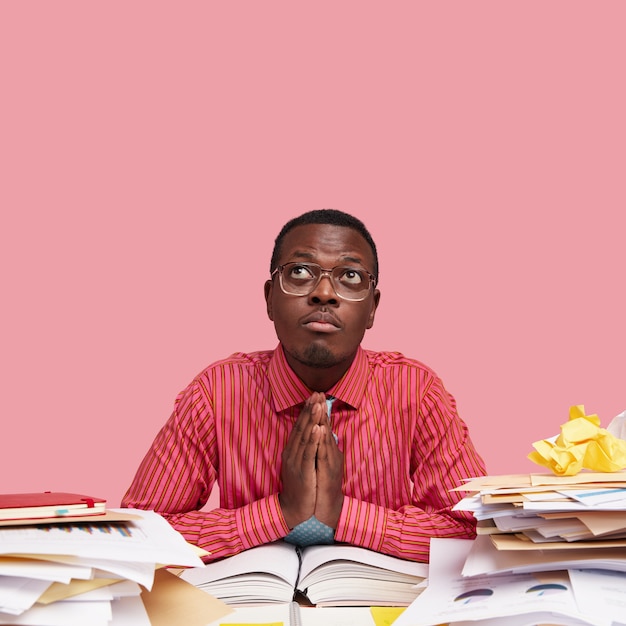 Dark skinned African American male prays for good luck, models over pink studio wall, keeps palms pressed together