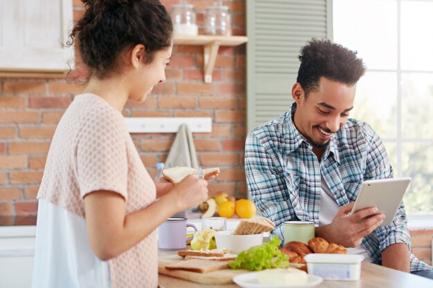 Dark skinned Afro American man dressed casually sits at kitchen with tablet computer, reads news online when his wife makes sandwhiches.
