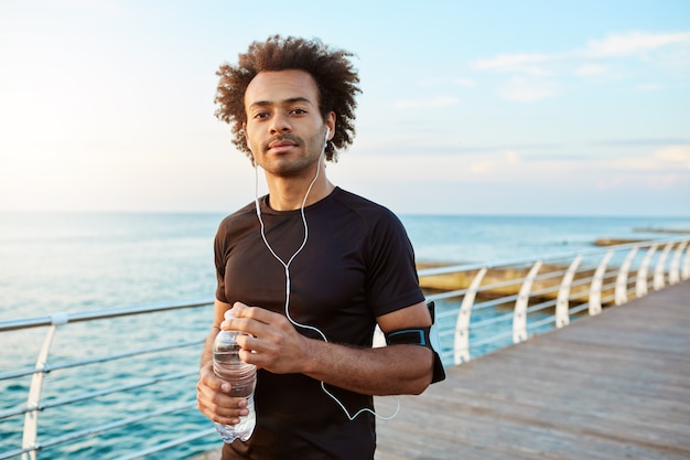 Dark-skinned cheerful sportsman drinking water out of plastic bottle, wearing earphones taking break during jogging. Portrait of dark-skinned athlete enjoying morning and music.