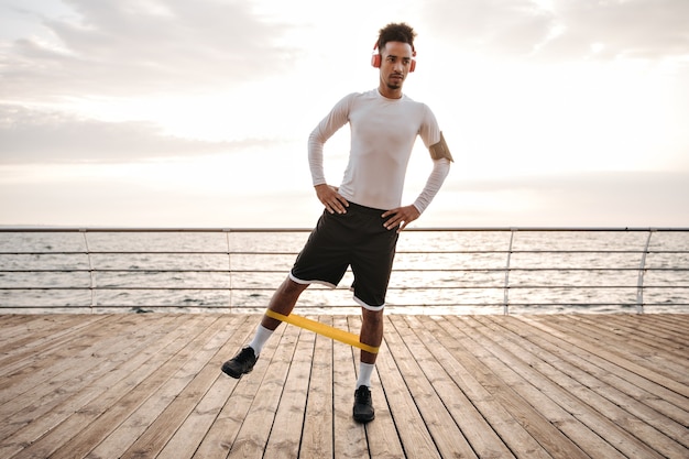 Dark-skinned curly brunette man in white long-sleeved t-shirt and black shorts listens to music in headphones and does exercises with fitness rubber near sea